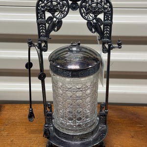 May include: An antique silver-plated butter dish with a clear, patterned glass jar. The dish has an ornate, decorative frame with a handle and a small serving utensil. The design includes floral and scrollwork details, and it sits on a three-legged base.