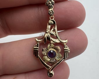 May include: A gold pendant with a purple gemstone in the center of a flower design. The pendant is surrounded by a delicate floral pattern and hangs from a chain with two small flowers.
