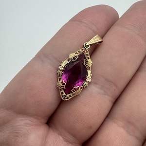 May include: A marquise-cut magenta gemstone pendant set in a yellow gold filigree frame. The metalwork features intricate fan-like patterns and delicate floral motifs surrounding the central stone, topped with a simple gold bail for hanging.