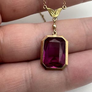 May include: A gold necklace featuring a rectangular pendant with a deep magenta gemstone. The pendant is suspended from a delicate gold chain by a small gold leaf-shaped connector and a tiny round bead, creating a refined and elegant jewelry piece.