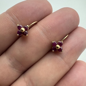 May include: A pair of gold earrings featuring a flower-shaped design. Each earring consists of four round, deep red rubies arranged as petals around a central gold bead, attached to a curved gold wire hook for secure placement in the earlobe.