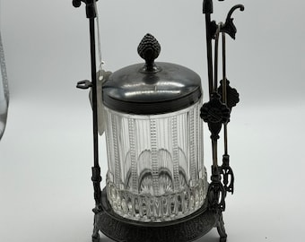 May include: A silver-plated glass sugar dispenser with a decorative metal stand. The stand features an ornate design with a curved arch and intricate details. The glass jar has a clear, ribbed design and a silver-plated lid with a finial.