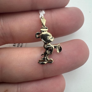 K&ouml;nnte beinhalten: Ein kleiner, goldfarbener Mickey Mouse Anh&auml;nger. Der Anh&auml;nger ist an einer d&uuml;nnen, wei&szlig;en Kordel befestigt. Die Mickey Mouse Figur befindet sich in einer klassischen Pose mit ausgestreckten Armen. Der Anh&auml;nger ist sehr detailliert.