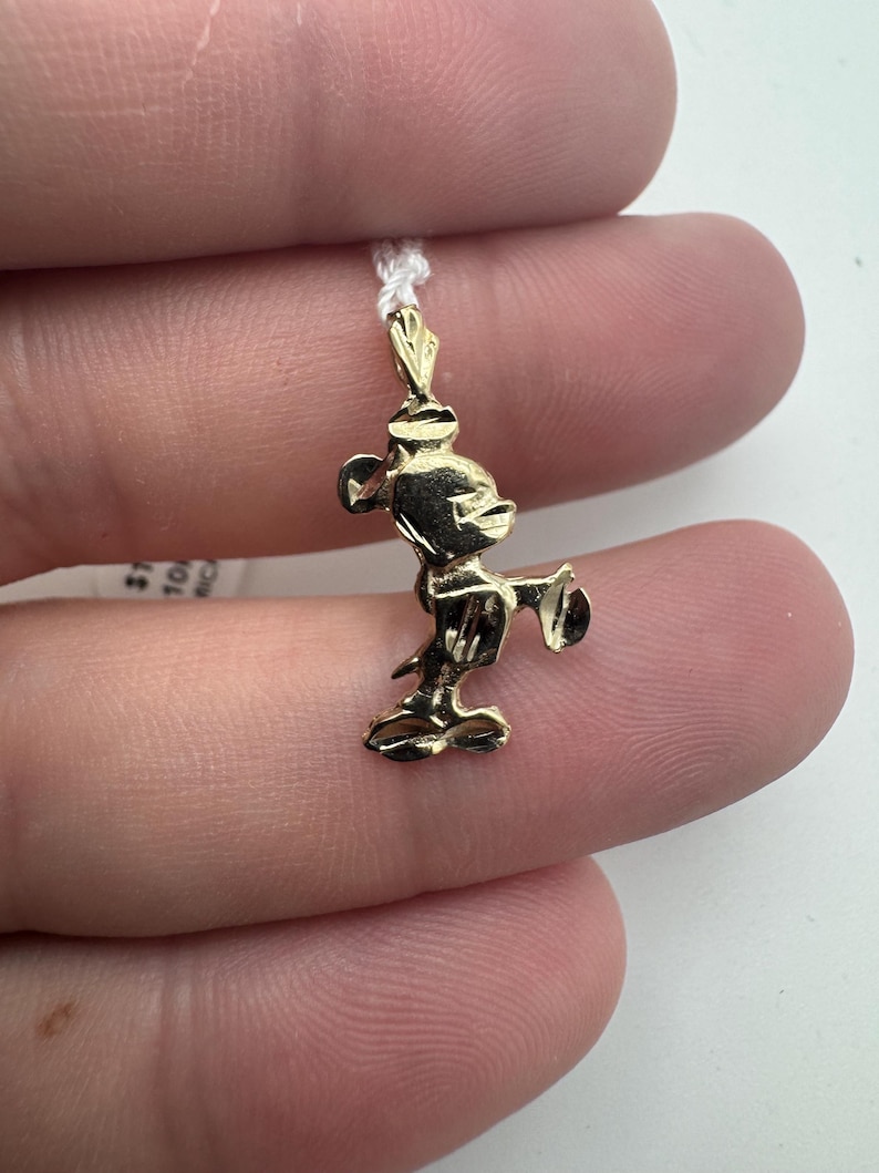 K&ouml;nnte beinhalten: Ein kleiner, goldfarbener Mickey Mouse Anh&auml;nger. Der Anh&auml;nger ist an einer d&uuml;nnen, wei&szlig;en Kordel befestigt. Die Mickey Mouse Figur befindet sich in einer klassischen Pose mit ausgestreckten Armen. Der Anh&auml;nger ist sehr detailliert.