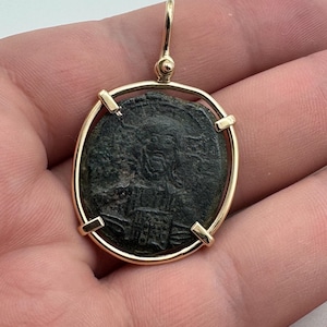 May include: A gold pendant featuring an ancient coin with a detailed relief of a figure. The coin is set within a gold frame with a loop for a chain. The coin's surface is dark with visible signs of age.