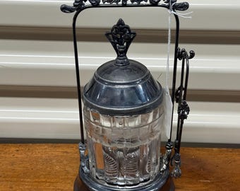May include: A silver-plated glass sugar dispenser with a decorative metal frame and a glass jar with a clear glass lid. The dispenser has a handle and a small spoon.