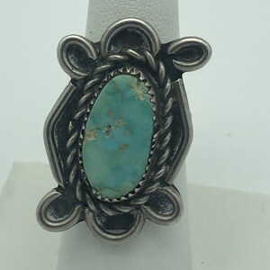 May include: A silver ring featuring a turquoise stone. The oval-shaped turquoise is set in a decorative frame with a twisted rope border. The ring has a detailed, ornate design with a vintage aesthetic.