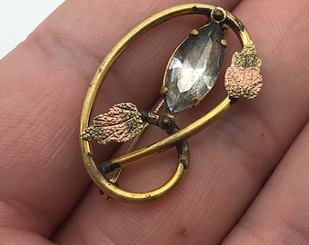 May include: A gold-toned brooch with a clear faceted stone set in a stylized oval frame. The frame is decorated with two gold-toned leaves with pink and green enamel.