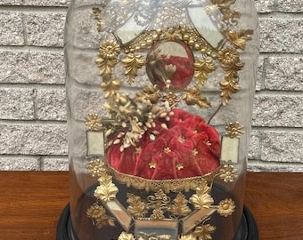 May include: A decorative glass dome with a black base, showcasing a red velvet cushion adorned with gold-colored embellishments. The dome features ornate gold accents, including floral and leaf designs, and a small mirror. The overall aesthetic is vintage and ornate.