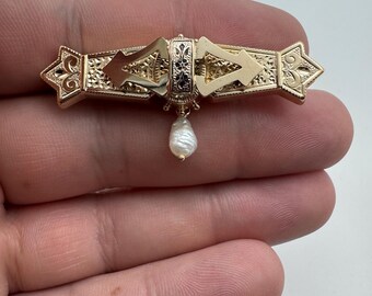 May include: A gold-toned brooch with intricate filigree and geometric designs. The brooch features a central rectangular element with black accents and a dangling teardrop-shaped pearl. The ends of the brooch have decorative arrow-like shapes.