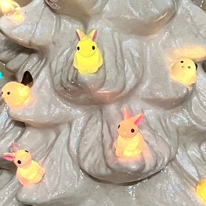 May include: A white ceramic Christmas tree with glitter accents, adorned with colorful lights and small bunny and bird figurines. The tree is lit with warm-toned bulbs and has a classic, festive design.