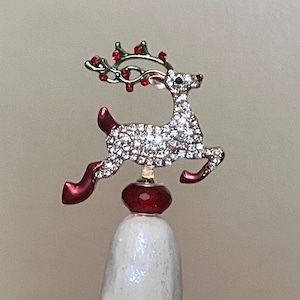 May include: A white ceramic Christmas tree with colorful lights. The tree is topped with a rhinestone reindeer figurine with red accents. The lights are red, blue, green, and yellow.