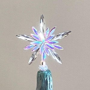 May include: A clear, iridescent star tree topper with multiple points. The star sits atop a green ceramic Christmas tree with red, yellow, and blue lights. The tree has a glossy finish and a classic design.