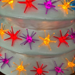 May include: A white ceramic Christmas tree with multi-colored star-shaped lights. The lights are in shades of red, orange, yellow, purple, and blue.