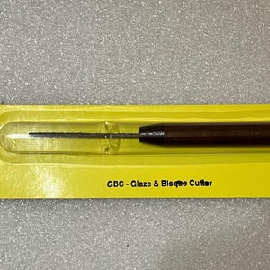 May include: A Kemper GBC Glaze & Bisque Cutter tool with a thin, serrated metal blade and a dark brown wooden handle, encased in a yellow blister pack with black text that reads Kemper and GBC - Glaze & Bisque Cutter, designed for ceramic work.