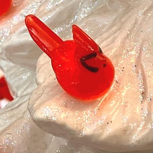 May include: A small, bright red cardinal ornament with black accents on its face. The bird is perched on a white, glittery surface, creating a festive and decorative appearance. The ornament is likely for holiday or seasonal decor.