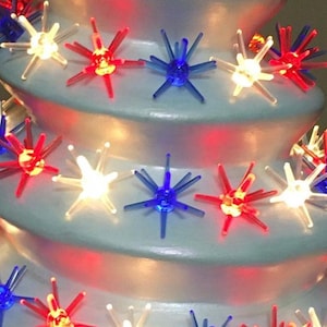 May include: A white ceramic Christmas tree with red, white, and blue star-shaped lights. The tree is decorated with a string of lights that are arranged in a spiral pattern.