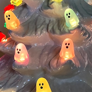 May include: A close-up of a ceramic Christmas tree with colorful ghost-shaped lights. The tree is a light blue color, and the ghost lights are in various colors, including yellow, orange, and pink. The lights have black dot eyes and are lit up.