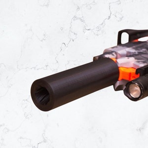 Puede incluir: Una extensión de cañón de plástico negro para una pistola de juguete. La extensión de cañón está unida a una pistola de juguete con un patrón de camuflaje. La pistola de juguete también tiene una punta de seguridad naranja y una linterna negra adjunta.