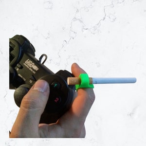 May include: A green finger holder for a cigarette, with a cigarette in it. The holder is being used by a person holding a black video game controller.