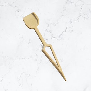 May include: A wooden craft stick with a spade-shaped end. The stick is light brown and has a geometric design.