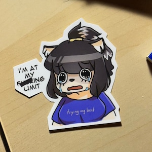 May include: A sticker featuring a cartoon character with black hair, blue eyes, and a blue shirt. The character is crying and has a speech bubble that says "I'm at my f***ing limit" with the word "f***ing" crossed out. The character is also wearing a blue shirt with the text "trying my best" on it.