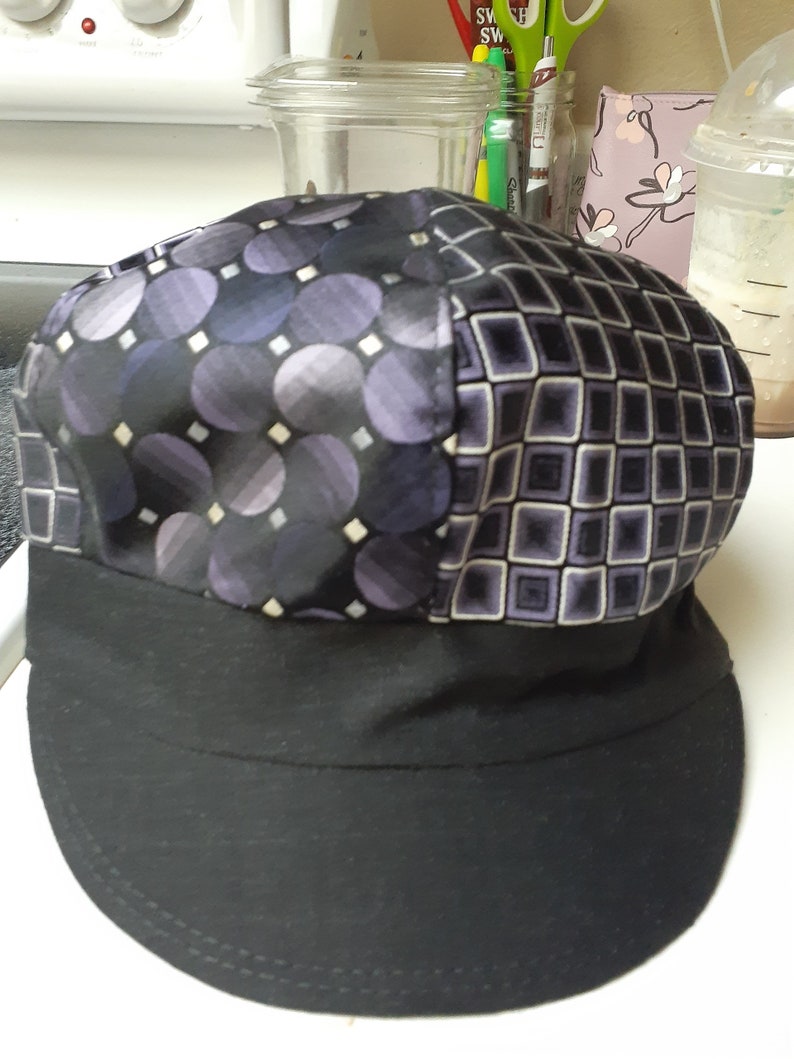 K&ouml;nnte beinhalten: Ein schwarzer Hut aus Stoff mit violettem Muster und schwarzem Rand. Der Hut hat ein geometrisches Muster mit Kreisen und Quadraten.