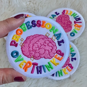 Peut inclure: Écussons brodés ronds avec l'inscription "PROFESSIONAL OVERTHINKER" en couleurs arc-en-ciel autour d'un cerveau rose. Les écussons ont un fond blanc et une bordure blanche.