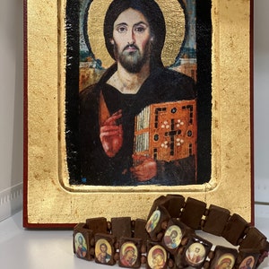 May include: Two brown wooden bracelets with small images of religious figures. The bracelets are on a white surface in front of a framed painting of Jesus.