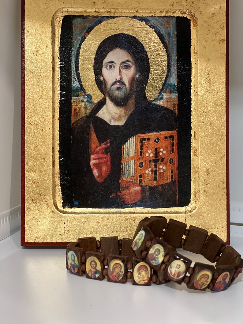 May include: Two brown wooden bracelets with small images of religious figures. The bracelets are on a white surface in front of a framed painting of Jesus.