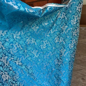 May include: A roll of turquoise fabric with a repeating pattern of silver dragons and clouds. The fabric has a shiny, textured appearance and is likely a decorative textile for clothing or home decor.