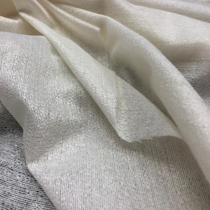 May include: Close-up of a cream-coloured fabric with a soft, textured appearance. The material is draped, showing folds and subtle variations in light and shadow. The fabric has a delicate, almost sheer quality.