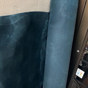 May include: A large roll of dark teal fabric with a matte, slightly textured surface. The material is tightly wound into a cylindrical shape, with a portion of the fabric unrolled and draped to the left, showing a consistent deep blue-green colour.