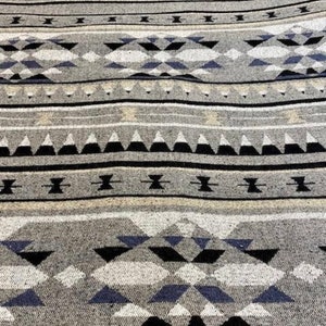 May include: Close-up of a grey textile with a geometric pattern. The design features repeating shapes in black, white, and blue, arranged in horizontal bands. The fabric has a textured appearance, suggesting a woven material.