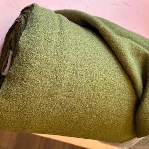 May include: A roll of olive green fabric with a textured, slightly fuzzy surface. The material is wound tightly, showing a dense weave and a consistent, earthy green color throughout the visible portion of the textile roll.
