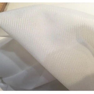 May include: Close-up of white fabric with a textured, woven pattern. The fabric is folded, creating shadows and highlighting the texture. The material appears lightweight, possibly for clothing or home decor.