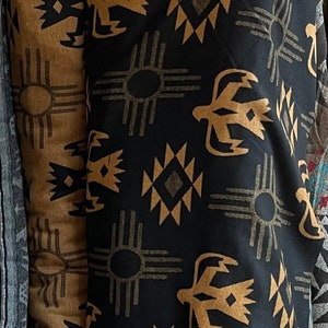 Peut inclure: Tissu noir avec un motif répétitif de formes géométriques brunes, y compris un symbole solaire et un oiseau stylisé. Le tissu a une texture douce et duveteuse.