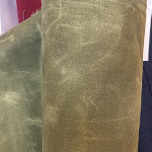 May include: A roll of olive green waxed canvas fabric featuring a textured, weathered appearance with visible creases and a frayed, raw edge at the top. The material displays a matte finish with subtle variations in colour depth across the surface.