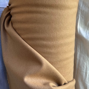May include: A roll of tan fabric, tightly wound. The fabric has a soft, matte texture and a warm, neutral color. The roll is partially unrolled, revealing the inner layers of the material. The fabric appears to be a medium weight, suitable for various sewing projects.
