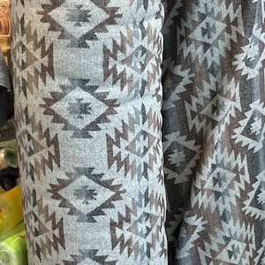 May include: Two rolls of fabric with a geometric pattern in shades of grey, brown, and black. The fabric has a woven texture and a fringed edge.