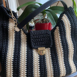 May include: A black and cream striped crochet handbag with a black leather handle and a small black flap with a gold clasp. The bag has a gold chain and clasp for attaching a shoulder strap.