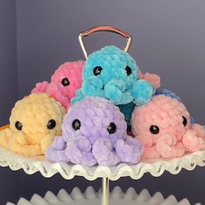 Könnte beinhalten: Fünf gehäkelte Tintenfisch-Plüschtiere in verschiedenen Farben, darunter rosa, blau, gelb und lila, werden auf einem weißen Kuchenständer ausgestellt. Jeder Tintenfisch hat schwarze Augen und ein strukturiertes, flauschiges Aussehen.