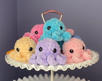 May include: Five crocheted octopus plushies in various colors, including pink, blue, yellow, and purple, are displayed on a white cake stand. Each octopus has black eyes and a textured, fluffy appearance.