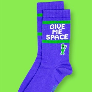 May include: A pair of purple socks with green detailing and the white text "GIVE ME SPACE". A green alien graphic is featured below the text. The socks have a green stripe at the top, set against a bright green background.