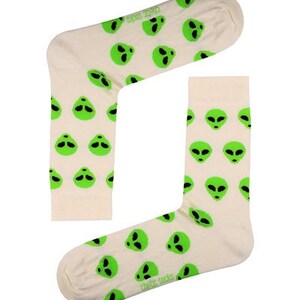 May include: A pair of cream-coloured socks featuring a repeating pattern of green alien faces. The socks have a ribbed cuff and the text "thebr socks" is printed on the sole. The socks are made of a soft, comfortable material.