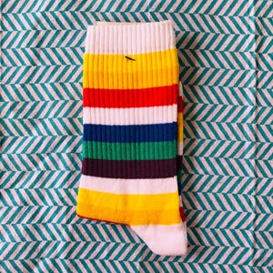 May include: A multi-coloured striped sock featuring bands of yellow, red, white, blue, green, and purple. The sock has a white cuff and toe, and is displayed on a teal and white chevron patterned fabric.
