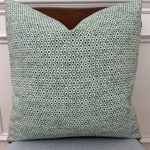 May include: A square throw pillow featuring a repeating geometric pattern of small dark blue dots centered within white diamond shapes, all set against a textured sage green background. The fabric has a visible woven weave and a plump, filled shape.