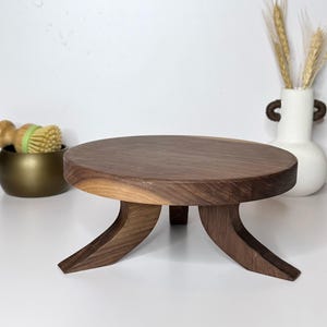 May include: A round, wooden pedestal with a dark brown finish. The pedestal has three curved legs and a flat top. The pedestal is approximately 8 inches in diameter and is suitable for displaying decorative items.