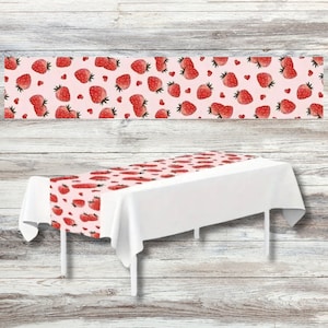 May include: A strawberry-themed table runner with a white tablecloth. The runner features a repeating pattern of red strawberries and small red hearts on a light pink background. The table runner is displayed on a white table.