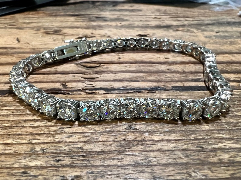 K&ouml;nnte beinhalten: Ein silbernes Tennisarmband mit einer Reihe von runden, klaren Edelsteinen. Das Armband liegt auf einer Holzoberfl&auml;che. Der Verschluss ist sichtbar, und das Armband scheint aus Metall zu sein.